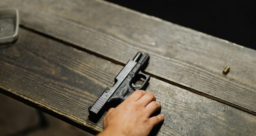Close-up of a hand holding a handgun on a wooden table with bullet casing.